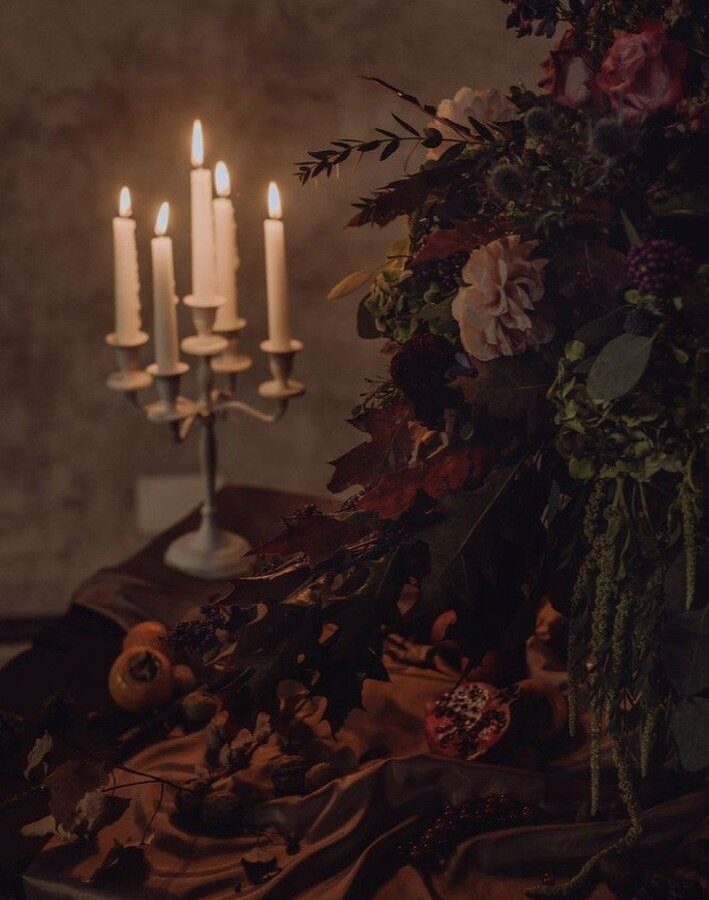 Candlelit candelabra surrounded by wilted roses, dried foliage, and autumn leaves on brown fabric in moody lighting.