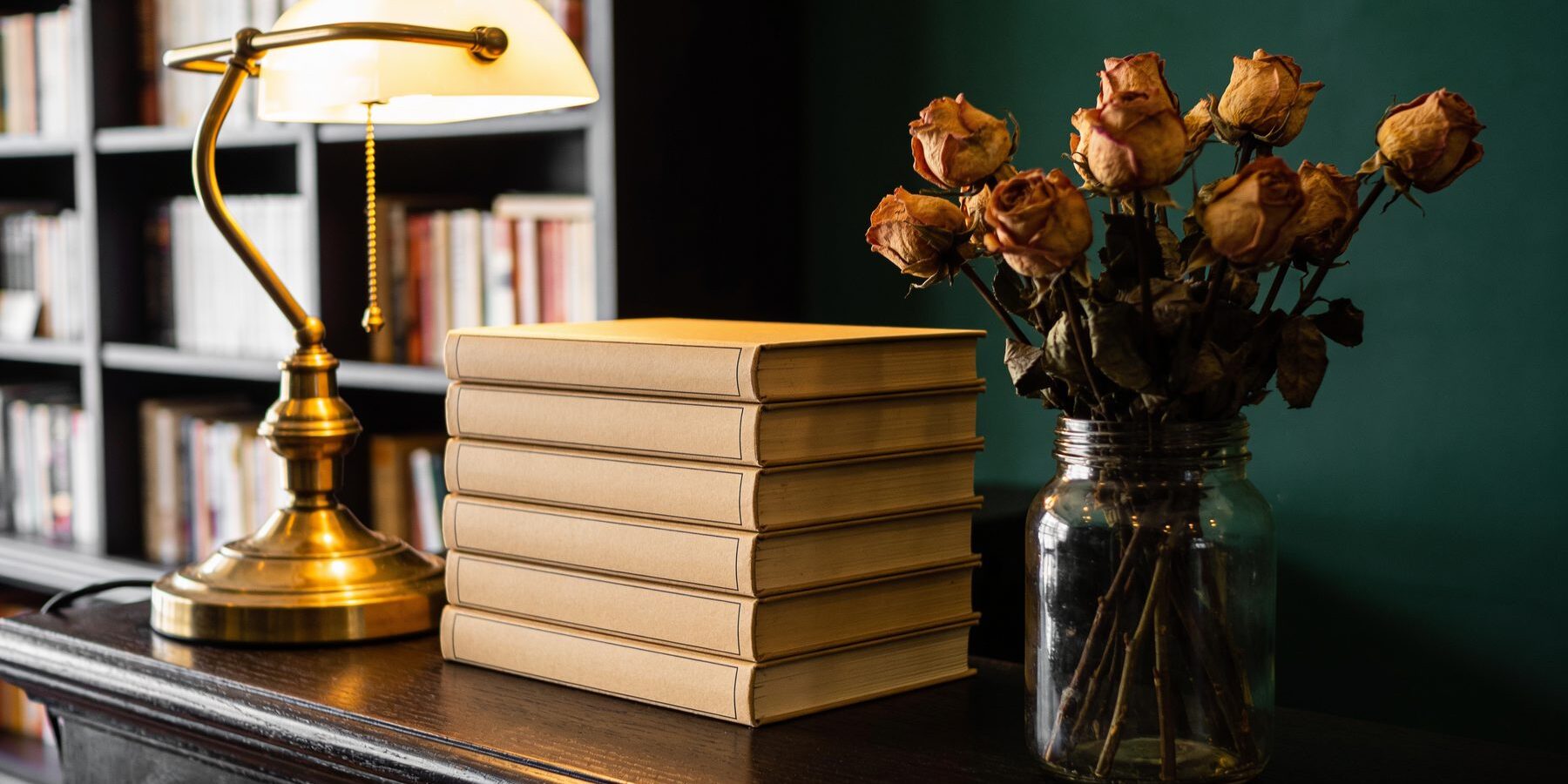 The shop counter in dark wood, a brass lamp, a stack of books and a jar of dried roses against a deep green wall.