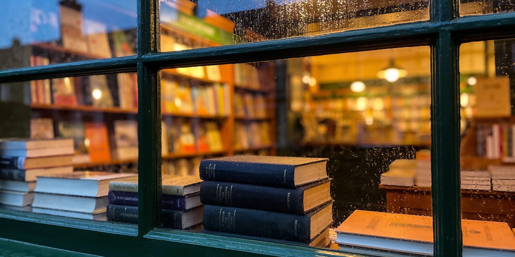 Rain on the shop window at dusk, stacked books on the sill and warm lamplight glowing from inside.