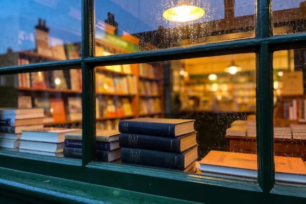 Rain on the shop window at dusk, stacked books on the sill and warm lamplight glowing from inside.